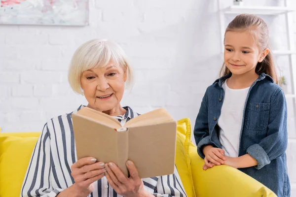 Lächelndes Kind steht neben Großmutter und liest Buch auf Couch — Stockfoto