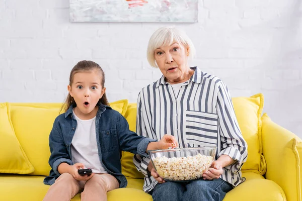 Mujer mayor asombrada sosteniendo tazón de palomitas de maíz cerca de niño con controlador remoto — Stock Photo