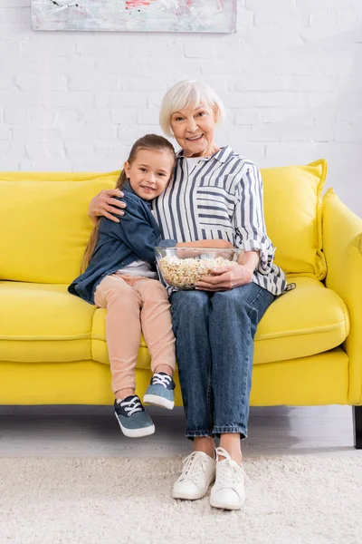 Bambino sorridente alla macchina fotografica mentre abbraccia la nonna con popcorn sul divano — Foto stock