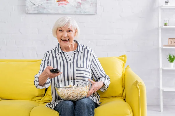 Mujer mayor positiva sosteniendo mando a distancia y palomitas de maíz en casa - foto de stock