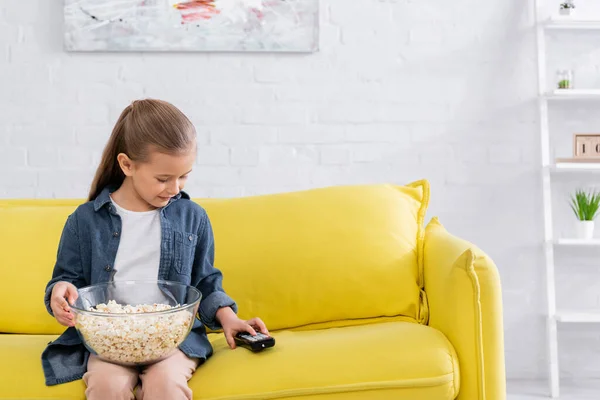 Girl holding bowl of popcorn and using remote controller on couch — Stock Photo