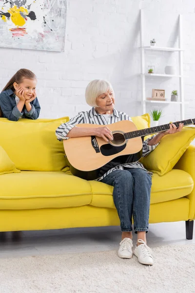 Nieta mirando a la abuela mayor tocando la guitarra acústica - foto de stock