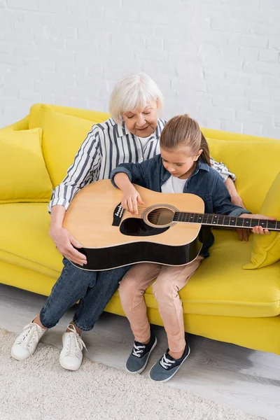 Criança tocando guitarra acústica enquanto avó sentada ao lado no sofá — Fotografia de Stock