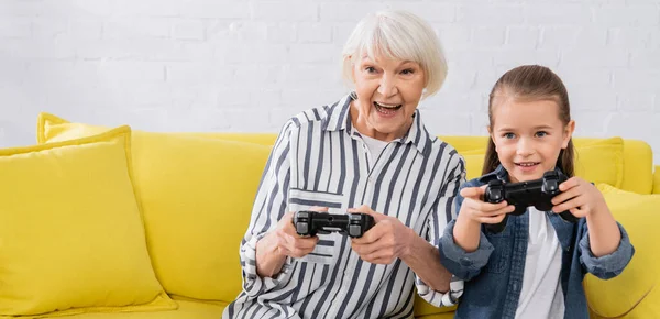 KYIV, UCRAINA - 11 GENNAIO 2021: nonna positiva che gioca ai videogiochi vicino alla nipote con joystick, striscione — Foto stock