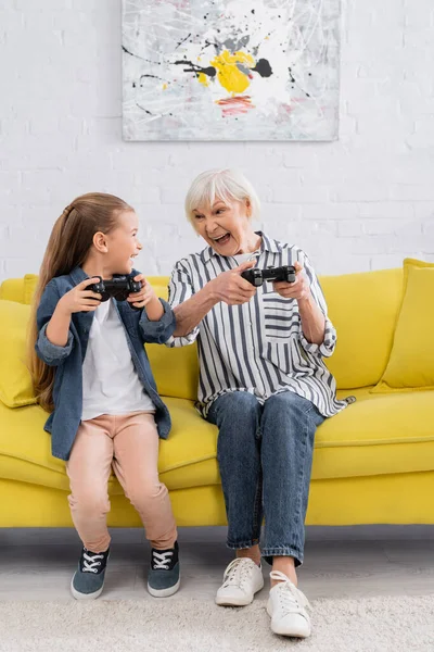 KYIV, UKRAINE - 11 JANVIER 2021 : Enfant excité avec joystick regardant grand-mère sur le canapé — Photo de stock