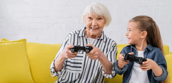 KYIV, UCRANIA - 11 de enero de 2021: Nieta sonriente con joystick mirando a la abuela en casa, pancarta - foto de stock