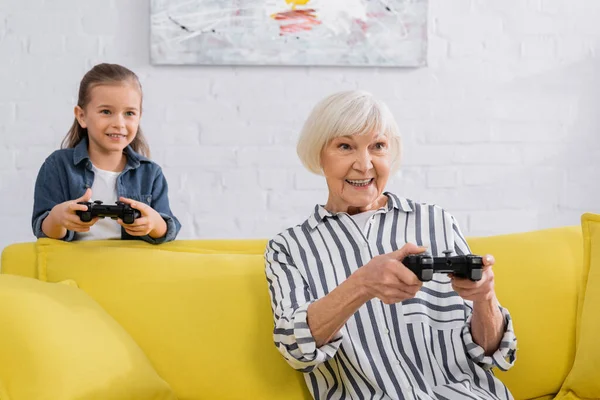 KYIV, UKRAINE - JANUARY 11, 2021: Smiling senior woman playing video game on couch near granddaughter — Stock Photo