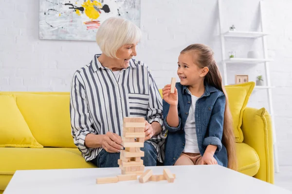 Улыбающаяся девушка держит блок деревянной игры рядом с бабушкой и башней на столе — стоковое фото