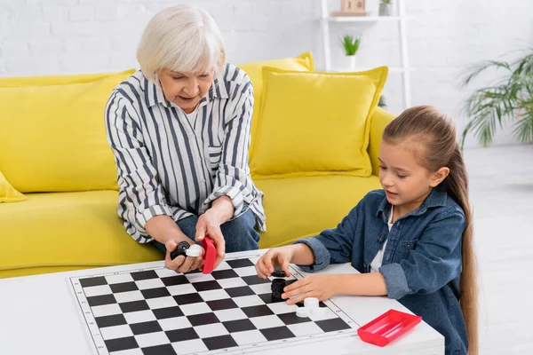 Oma hält Schachbrett neben Kind und Couchtisch im Wohnzimmer — Stockfoto