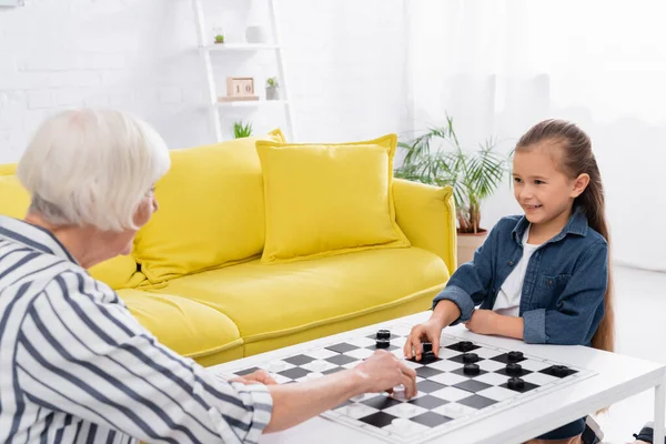 Smiling kid playing checkers with blurred grandmother at home — Stock Photo