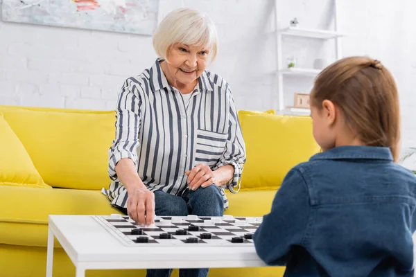 Femme âgée souriant tout en jouant aux dames avec enfant flou — Photo de stock