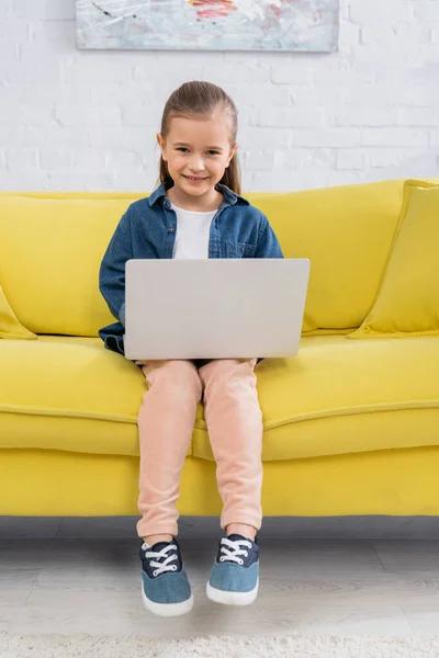 Criança com laptop sorrindo para a câmera no sofá amarelo — Fotografia de Stock