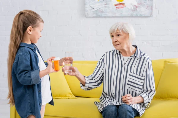Cuidado chica sosteniendo pastillas y agua cerca de la abuela en el sofá - foto de stock
