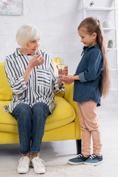 Senior woman taking glass of water from smiling granddaughter at home — Stock Photo