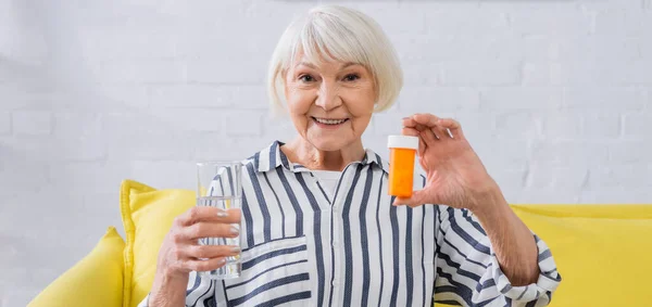 Mujer anciana sonriendo a la cámara mientras sostiene un vaso de agua y un frasco de píldoras, pancarta - foto de stock