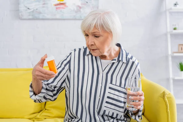 Mujer mayor mirando frasco con pastillas y sosteniendo vaso de agua en casa - foto de stock