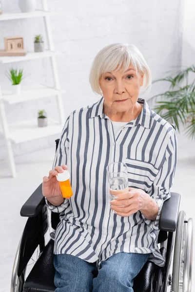 Femme âgée avec des pilules et de l'eau regardant la caméra en fauteuil roulant — Photo de stock