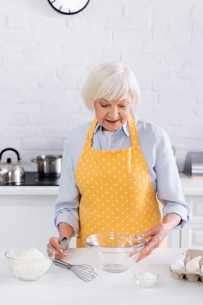 Sorridente donna anziana guardando gli ingredienti sul tavolo della cucina — Foto stock