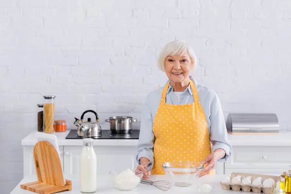 Sonriente anciana mirando a la cámara cerca de los ingredientes en la mesa de la cocina - foto de stock