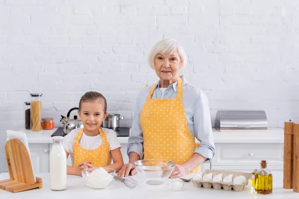 Grand-mère et enfant souriant regardant la caméra près des ingrédients et de la tablette numérique — Photo de stock
