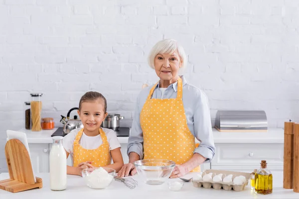 Abuela y niño sonriente mirando la cámara cerca de los ingredientes y la tableta digital - foto de stock