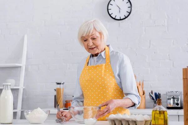 Mujer mayor en delantal tomando harina cerca de la leche y los huevos en la cocina — Stock Photo