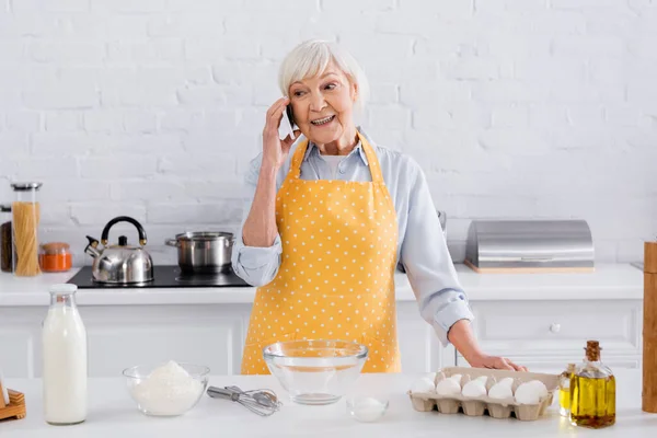 Donna anziana che parla sullo smartphone vicino agli ingredienti sul tavolo della cucina — Foto stock