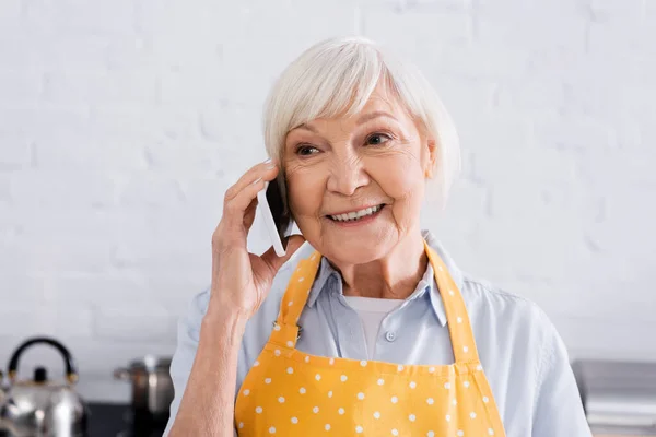 Lächelnde ältere Frau in Schürze unterhält sich in Küche mit Smartphone — Stockfoto