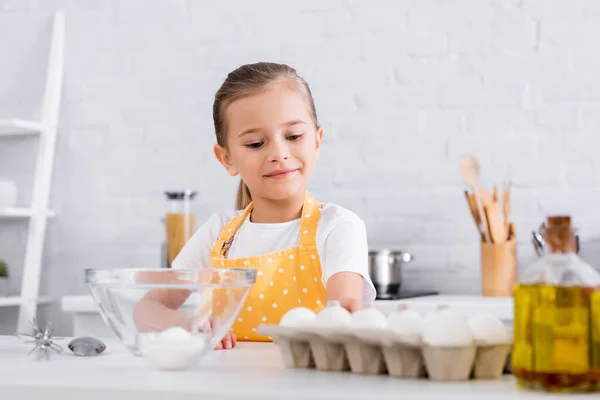 Kid in apron standing near blurred eggs and oil in kitchen — Stock Photo