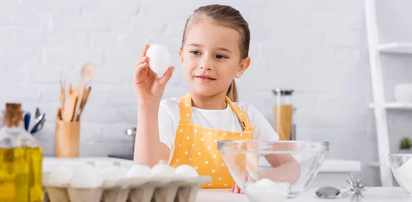 Chica en delantal sosteniendo huevo cerca de aceite y azúcar en la cocina, pancarta - foto de stock