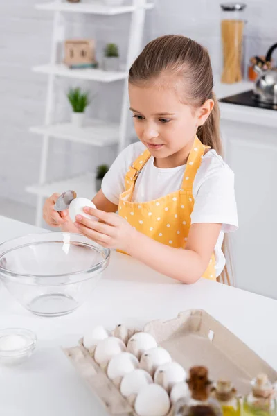 Enfant dans un tablier tenant une cuillère et un œuf près du sucre et de l'huile — Photo de stock