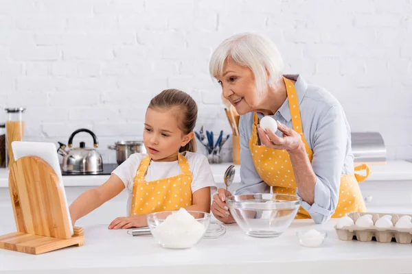 Sonriente abuelita y niño usando tableta digital cerca de los ingredientes en la cocina - foto de stock