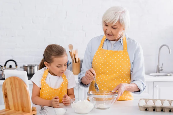Sonriente abuela sosteniendo cuchara cerca de harina, huevos y nieta en la cocina - foto de stock