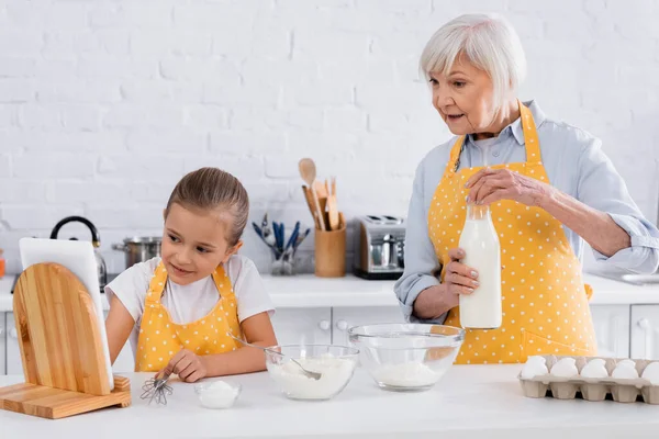 Ältere Frau hält Flasche Milch in der Nähe von Zutaten und Kind mit digitalem Tablet in Küche — Stockfoto