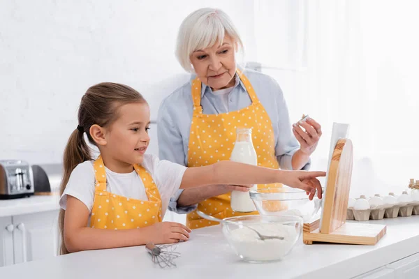 Alegre niño usando tableta digital cerca de ingredientes y abuela en casa - foto de stock