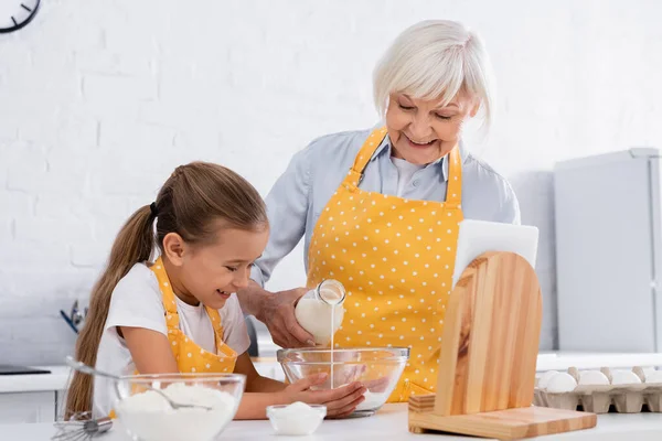 Positive kid and grandmother pouring milk in bowl near digital tablet and flour — Stock Photo