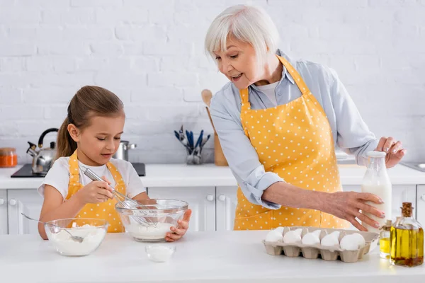 Sorrindo vovó segurando garrafa de leite perto de criança, ovos e farinha na cozinha — Fotografia de Stock