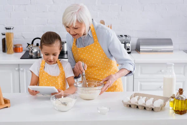 Ребенок с помощью цифрового планшета рядом с бабушкой приготовления пищи на кухне — стоковое фото