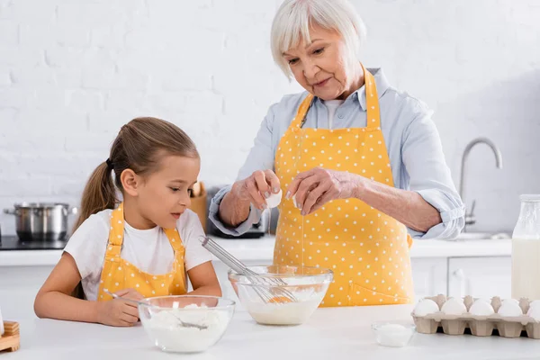 Mujer mayor vertiendo huevo en tazón cerca de los ingredientes y nieta en la cocina - foto de stock