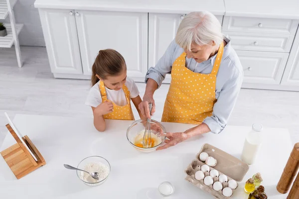 Vista aérea de la abuela mezclando huevos en un tazón cerca de la nieta y la tableta digital - foto de stock