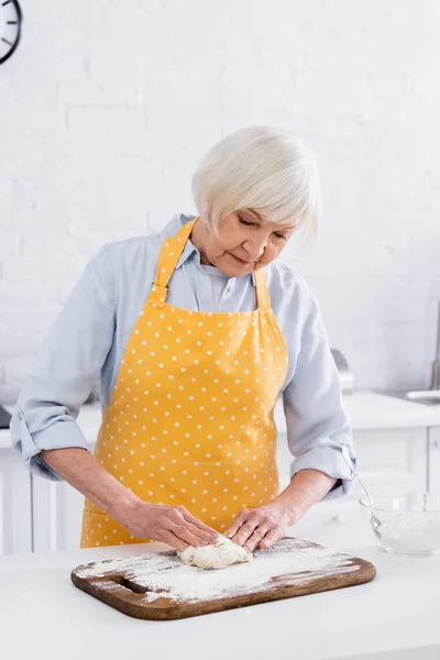 Elderly woman in apron crumpling dough on cutting board — Stock Photo