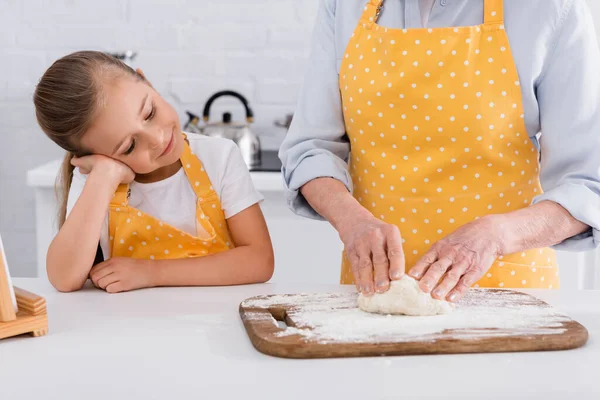 Kid in apron standing near granny making dough in kitchen — Stock Photo
