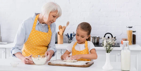 Ребенок делает тесто рядом с бабушкой в фартуке держа муку, плакат — стоковое фото