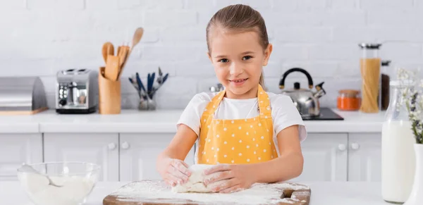 Улыбающийся ребенок в фартуке делает тесто рядом с молоком и мукой, плакат — стоковое фото