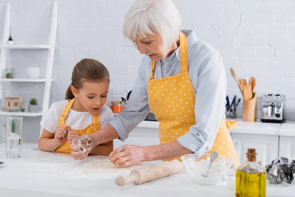 Abuela y nieta de pie cerca de la masa y cortador de galletas en la cocina - foto de stock