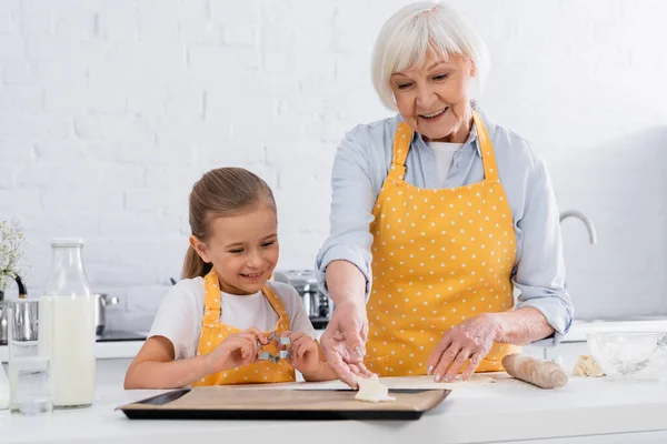 Улыбающийся ребенок держит нож для печенья рядом с бабушкой с тестом — стоковое фото