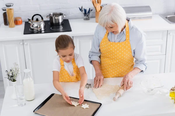 Vista ad alto angolo del bambino che mette la pasta sulla teglia vicino alla nonna con il mattarello — Foto stock