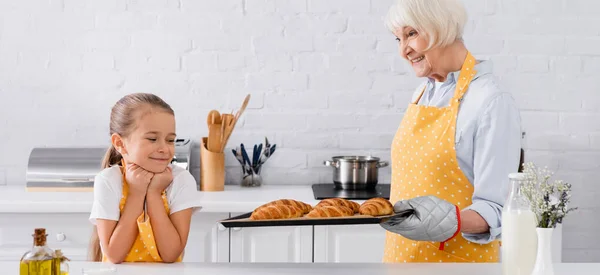 Grand-mère tenant des croissants sur une plaque à pâtisserie près d'un enfant souriant, bannière — Photo de stock