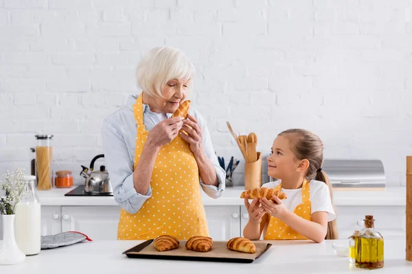 Abuela mayor olor croissant cerca de la nieta en la cocina - foto de stock