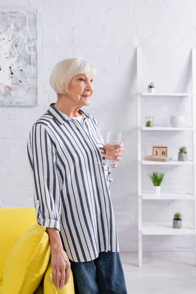 Улыбающаяся пожилая женщина держит стакан воды возле дивана — стоковое фото